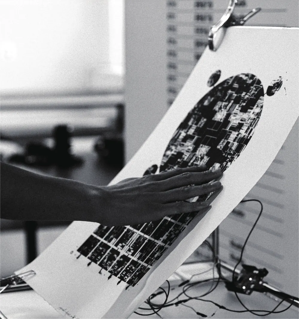 A hand pressing on a silkscreen poster clamped upright to a stand, with a printed circuit-like graphic across it.