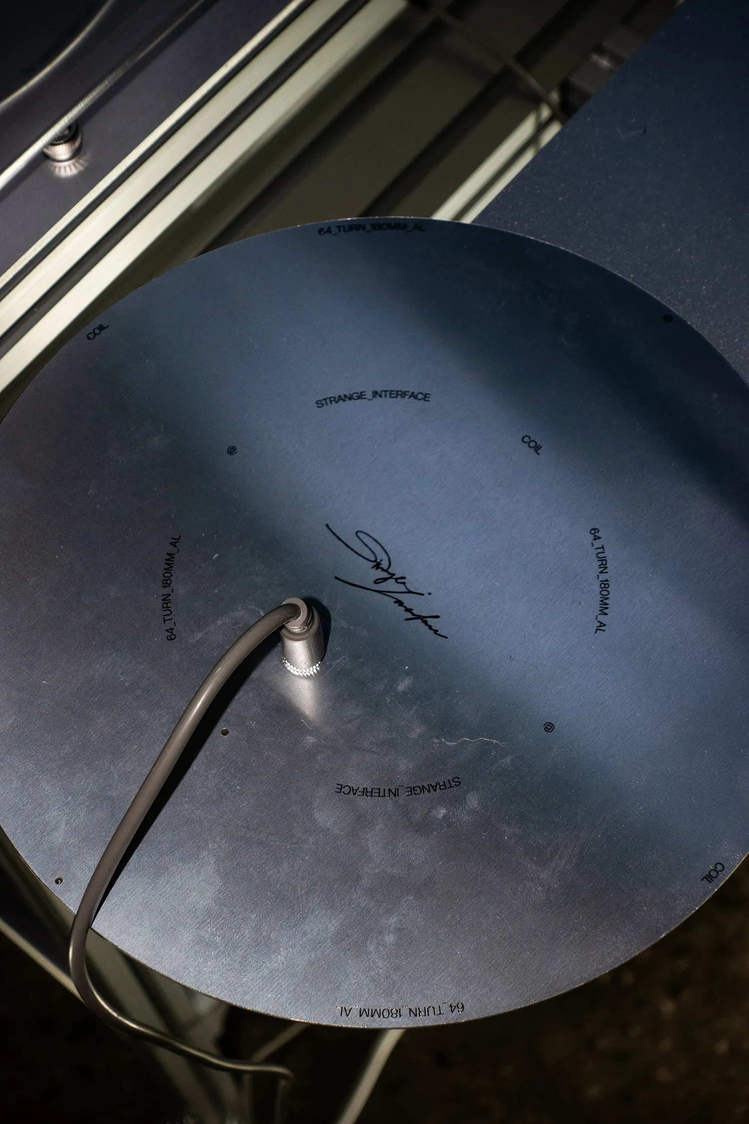 An aluminium disc engraved with the words COIL, STRANGE_INTERFACE and 64_TURN_180MM_AL around its rim, with a signature at the centre and a cable entering it.