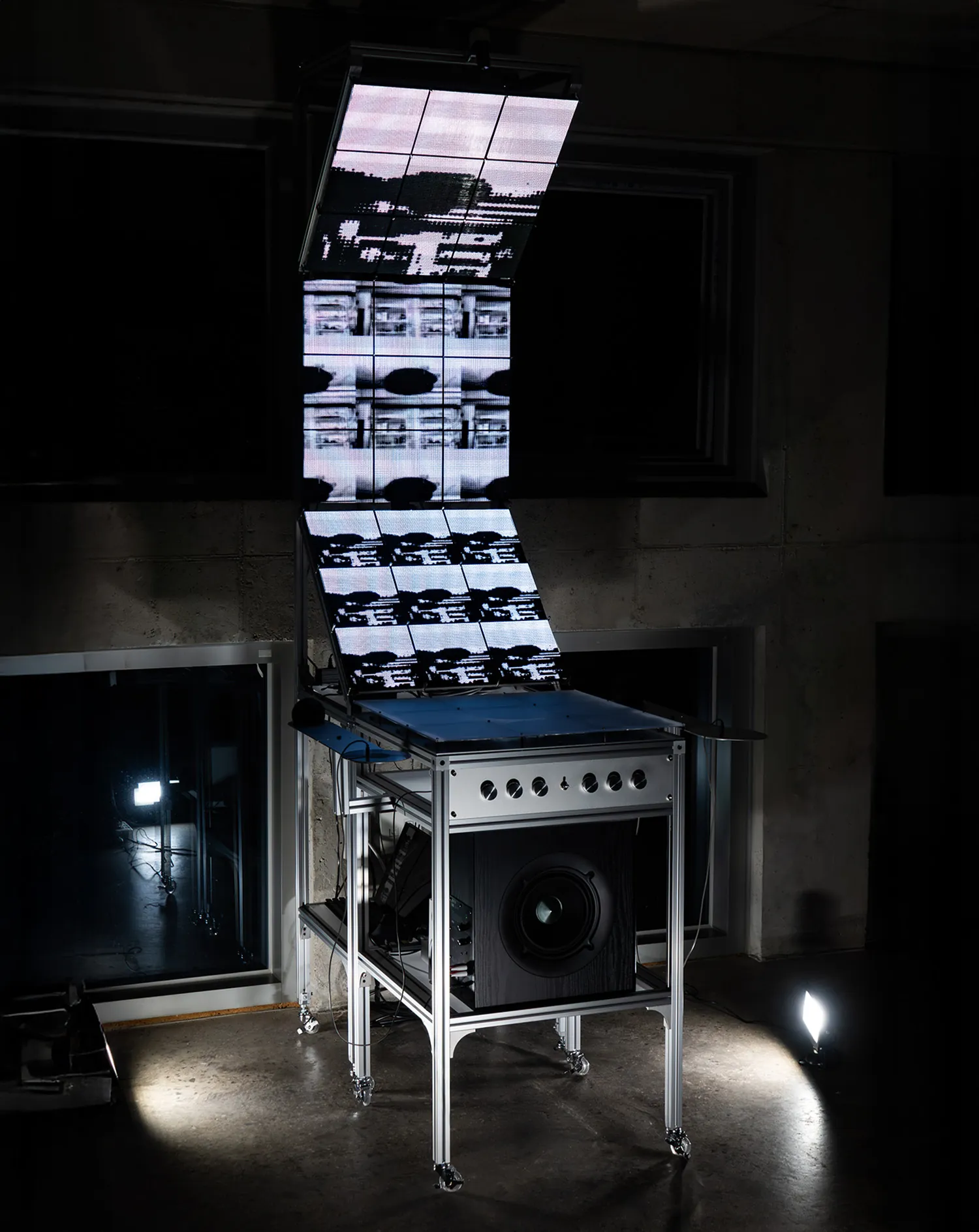 The whole rig lit in a dark room: a folded stack of LED panels rises from a wheeled aluminium cabinet with a row of knobs and a speaker.